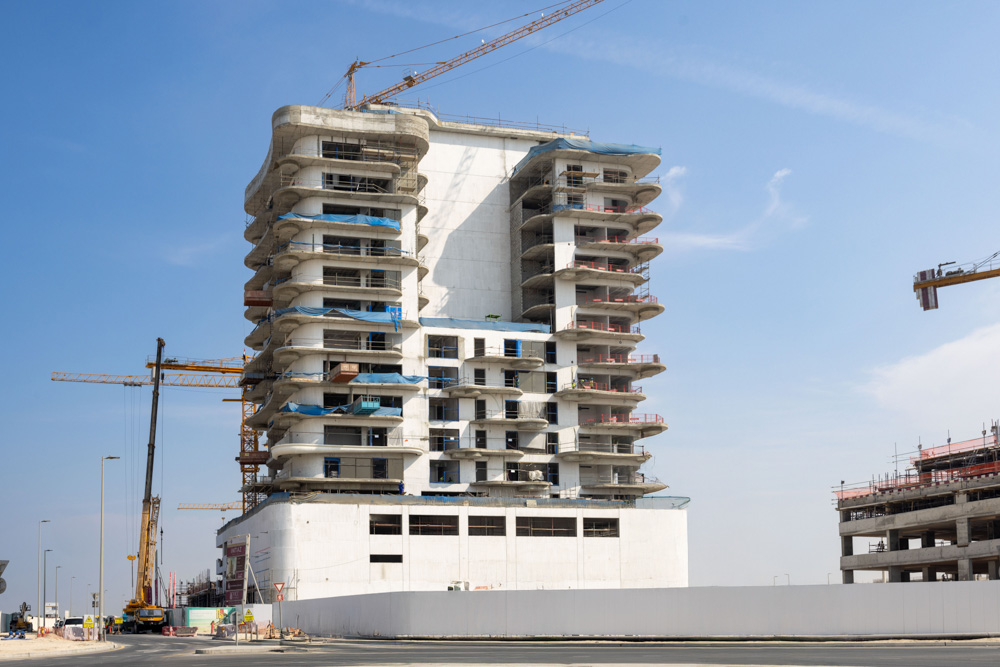 Beach Walk Iii By Imtiaz A partially constructed high-rise building with a crane, showcasing open balconies and a clear blue sky.